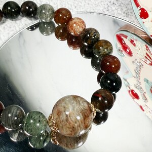 May include: A beaded bracelet featuring a variety of earth-toned gemstones. The beads range in color from deep green and black to reddish-brown and speckled tan. The bracelet is displayed on a reflective surface, showcasing its polished appearance.
