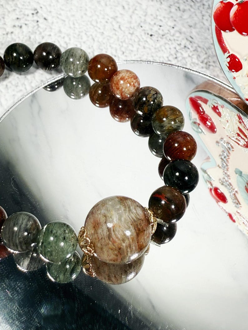 May include: A beaded bracelet featuring a variety of earth-toned gemstones. The beads range in color from deep green and black to reddish-brown and speckled tan. The bracelet is displayed on a reflective surface, showcasing its polished appearance.
