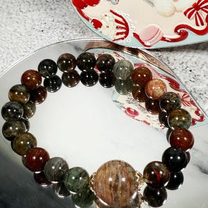 May include: A beaded bracelet featuring a large, central bead and smaller beads in shades of brown, green, and black. The bracelet is displayed on a reflective surface, with a decorative tin in the background.
