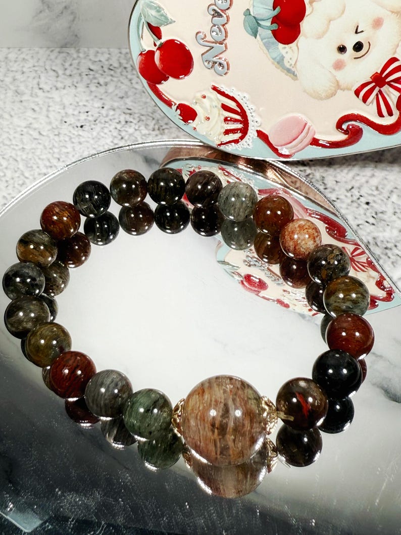 May include: A beaded bracelet featuring a large, central bead and smaller beads in shades of brown, green, and black. The bracelet is displayed on a reflective surface, with a decorative tin in the background.