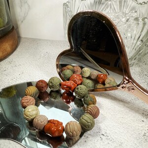 May include: A heart-shaped, rose gold hand mirror reflects a collection of colorful, textured beads. The beads are arranged on a silver, leaf-shaped tray. The beads are various colors, including brown, green, and orange.