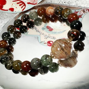 May include: A beaded bracelet featuring a variety of earth-toned beads, including brown, green, and black. The bracelet has a large, round, patterned bead as a focal point. The beads are strung together with a gold-colored chain.