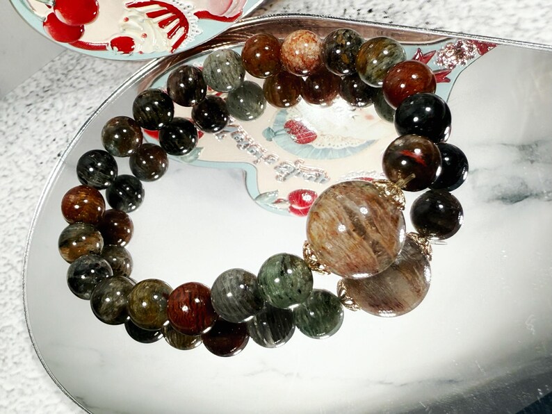 May include: A beaded bracelet featuring a variety of earth-toned beads, including brown, green, and black. The bracelet has a large, round, patterned bead as a focal point. The beads are strung together with a gold-colored chain.
