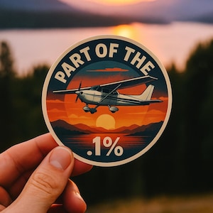 May include: A round sticker featuring a small airplane silhouetted against a sunset over a lake. The text "PART OF THE" arcs around the top, with ".1%" at the bottom. The sticker is held in a hand.