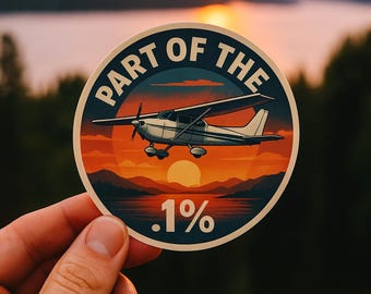 May include: A round sticker featuring a small airplane silhouetted against a sunset over a lake. The text "PART OF THE" arcs around the top, with ".1%" at the bottom. The sticker is held in a hand.
