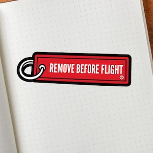 May include: A red rectangular tag with the text "REMOVE BEFORE FLIGHT" in white lettering, attached to a black and white keyring. The tag rests on an open, dotted notebook next to a white teacup and saucer with coffee and a silver pen.