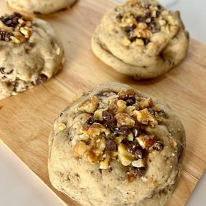 May include: Close-up of four round cookies on a wooden cutting board. The cookies are topped with chopped walnuts and chocolate chips.