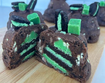 Puede incluir: Primer plano de una galleta brownie de chocolate con capas de galletas Oreo de menta. La galleta está cortada por la mitad, revelando las capas de galletas Oreo de menta en el interior. La galleta está sobre una tabla de cortar de madera.