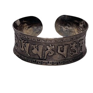 Puede incluir: Un brazalete plateado con escritura grabada. La banda ancha presenta un borde geométrico y el texto central está en una fuente estilizada. Los extremos abiertos del brazalete son redondeados. El brazalete se muestra sobre un fondo blanco.