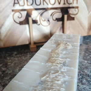 May include: A white bar of soap with a natural loofah embedded in it. The soap is on a wooden surface with the name "AJDUKOVICH" engraved on it.