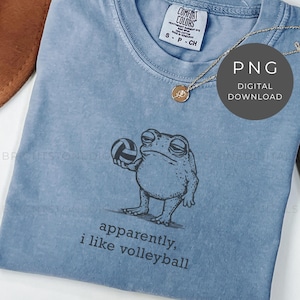 May include: A light blue t-shirt with a graphic of a frog holding a volleyball. The text "apparently, i like volleyball" is printed below the frog. A gold necklace with a pendant is on the shirt.