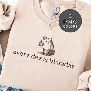 Peut inclure: Un sweat-shirt beige avec l'inscription "every day is blursday" en marron. Un dessin de grenouille tenant une tasse de café et une mallette est imprimé au-dessus du texte. Un graphique rond indique deux couleurs PNG disponibles.