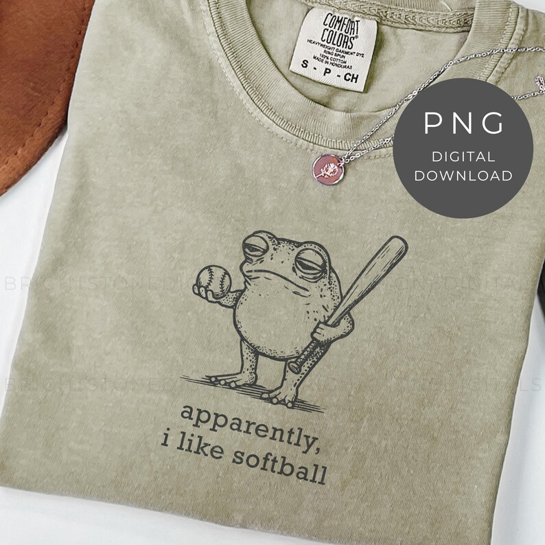 May include: A sage green t-shirt featuring a cartoon frog holding a baseball and bat. The text "apparently, i like softball" is printed below the frog. A silver necklace with a pink pendant is visible.