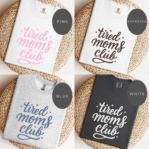 May include: Four t-shirts in white, espresso, blue, and black, each with the text "tired moms club" in different colors. The shirts are displayed on a woven, circular surface, with color labels: pink, espresso, blue, and white.