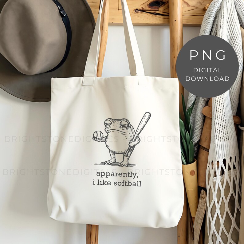 May include: Off-white tote bag featuring a cartoon frog holding a baseball and bat. The frog has a bored expression. Text on the bag reads "apparently, i like softball". The bag has long straps and is hanging on a wooden rack.