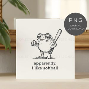 May include: A black and white illustration of a frog holding a baseball bat and ball. The text "apparently, i like softball" is below the frog. A digital download PNG graphic.