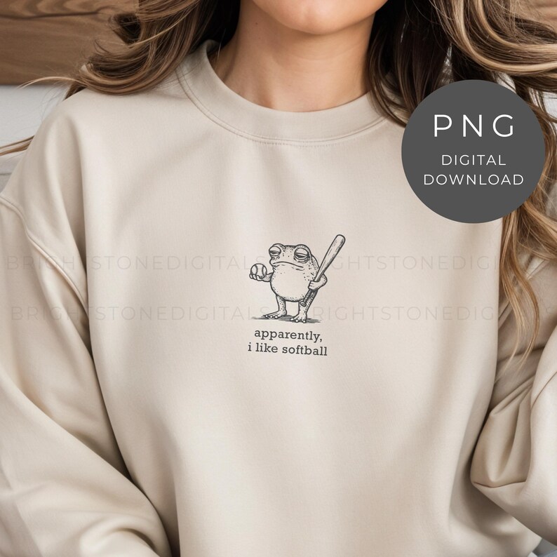 May include: Beige sweatshirt featuring a black and white cartoon frog holding a baseball bat and ball. The text "apparently, i like softball" is printed below the frog. The image is a digital download.