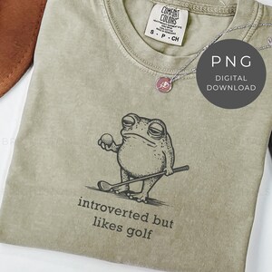 May include: A sage green t-shirt featuring a cartoon frog holding a golf ball and club. The text "introverted but likes golf" is printed below the frog. A silver necklace with a pink pendant is on the shirt. The shirt has a Comfort Colors tag.