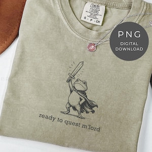 May include: A sage green t-shirt featuring a cartoon frog in a cape holding a sword aloft. The text "ready to quest m'lord" is printed below the frog. A silver necklace with a pink pendant is visible. The shirt has a Comfort Colors tag.