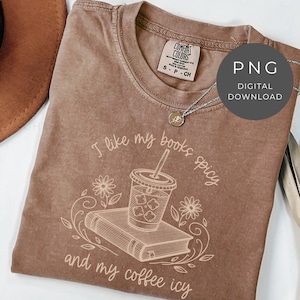 May include: A brown Comfort Colors t-shirt with the text "I like my books spicy and my coffee icy" in a cursive font. The graphic includes a book, iced coffee, and floral accents. A silver necklace and a brown hat are also visible.