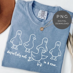 May include: A blue Comfort Colors t-shirt with a white graphic of five cartoon ducks. The text "definitely not in a row..." is below the ducks. A gold necklace and a brown hat are also visible.