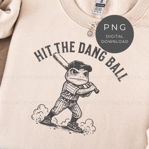 May include: A beige sweatshirt with the text "HIT THE DANG BALL" in black, featuring a cartoon frog in a baseball uniform. The frog is holding a bat, ready to swing. A digital download PNG graphic is also visible.