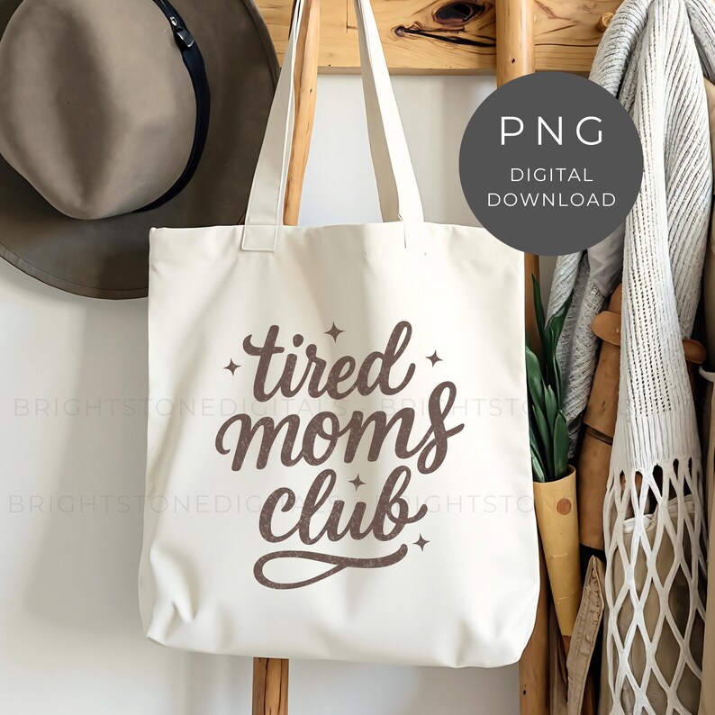 May include: Cream-colored tote bag with the text "tired moms club" in a brown script font. The bag has long straps and is hanging on a wooden rack. A hat and other items are visible in the background. A digital download PNG graphic is also visible.