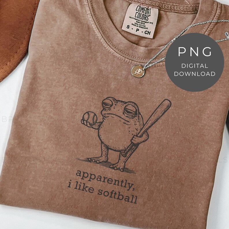 May include: Brown Comfort Colors t-shirt with a graphic of a frog holding a baseball and bat. The text "apparently, i like softball" is printed below the frog. A silver necklace and a digital download graphic are also visible.