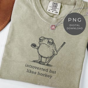 Puede incluir: Camiseta verde salvia con un dibujo de una rana jugando al hockey. El texto "introverted but likes hockey" está impreso debajo. También se ve un collar plateado con un colgante.