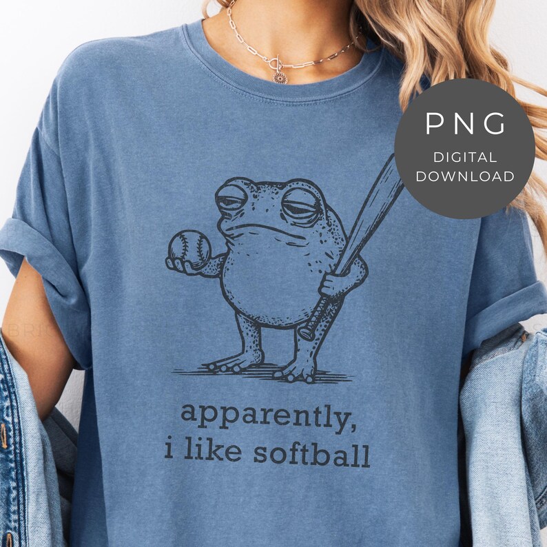 May include: A blue t-shirt featuring a cartoon frog holding a baseball bat and ball. The text "apparently, i like softball" is printed below the frog. A digital download PNG graphic is in the upper right corner.