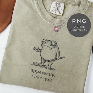 May include: A sage green t-shirt featuring a cartoon frog holding a golf ball and club. The text "apparently, i like golf" is printed below the frog. A silver necklace with a pink pendant is visible.