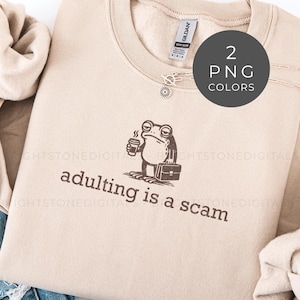 May include: Beige sweatshirt with the phrase "adulting is a scam" and a cartoon frog holding a coffee cup and briefcase. The sweatshirt has a crew neck and long sleeves. A small silver charm is attached to the neckline.