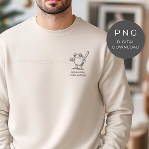 May include: Cream-colored sweatshirt featuring a small black and white illustration of a frog holding a baseball bat and ball. The text "apparently, I like softball" is below the frog.