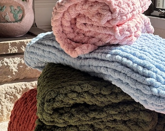 Puede incluir: Una pila de cuatro mantas de punto grueso con una construcción de hilo suave y texturizado. Las mantas están superpuestas en colores rosa claro, azul claro, verde oliva oscuro y naranja terracota, cada una con un patrón de puntada gruesa.