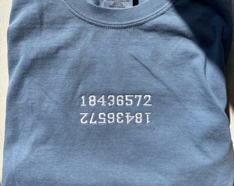 Puede incluir: Una camiseta de cuello redondo en color azul polvoriento con texto bordado en blanco que dice 18436572, con el mismo número repetido justo debajo en una orientación invertida y reflejada. La etiqueta interior del cuello muestra la marca Comfort Colors.