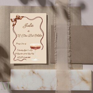Puede incluir: Una tarjeta de invitación beige con una cinta marrón y una copa de vino tinto. El texto de la tarjeta dice "Julie is A Tini Bit Older. Please join us Saturday April 3, at 7p 123 Rugastare street, May City RSVP 111-000-9999"
