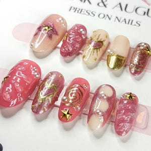 May include: Set of ten pink and gold press-on nails with various designs, including stars, swirls, and hearts.  "Pink & Aug" and "Press On Nails" are printed on the background.