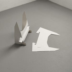 Puede incluir: Dos expositores de cartón blanco, uno montado y otro desmontado, están colocados sobre una superficie gris. El expositor montado tiene forma triangular con una balda. La pieza desmontada tiene una forma única con un gancho.