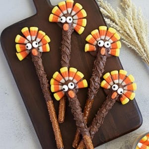 Puede incluir: Barritas de pretzel con temática de Acción de Gracias decoradas como pavos. Cada pavo tiene un cuerpo de barrita de pretzel cubierto de chocolate, plumas de caramelos de maíz, ojos de caramelo y un pico de caramelo naranja.