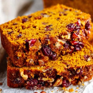 Könnte beinhalten: Zwei Scheiben Kürbisbrot mit sichtbaren Cranberries und Walnüssen. Das Brot hat eine goldorange Farbe und eine strukturierte Oberfläche, was auf ein feuchtes und schmackhaftes Backwerk hindeutet. Die Zutaten sind gleichmäßig verteilt.