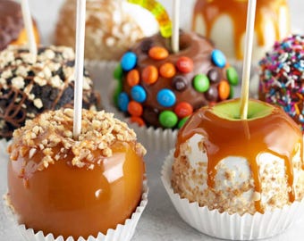 Op de afbeelding: Een verzameling karamelappels met diverse toppings, waaronder noten, chocolade, hagelslag en gekleurde chocoladesnoepjes. Elke appel zit op een houten stokje en wordt gepresenteerd in een wit papieren bakje.