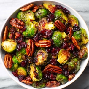 Può includere: Una ciotola bianca piena di cavoletti di Bruxelles arrostiti, noci pecan e mirtilli rossi secchi. I cavoletti sono verdi e dorati, con le noci e i mirtilli che aggiungono consistenza e colore. Sono visibili anche pezzi di pancetta.