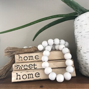 May include: A stack of three books with the words "home sweet home" written on them. The books are decorated with a white beaded garland and a jute tassel.