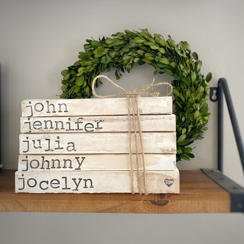 May include: A stack of five white books with names printed on the spines: "john", "jennifer", "julia", "johnny", and "jocelyn". The books are tied together with twine and a small heart is visible on the bottom book.