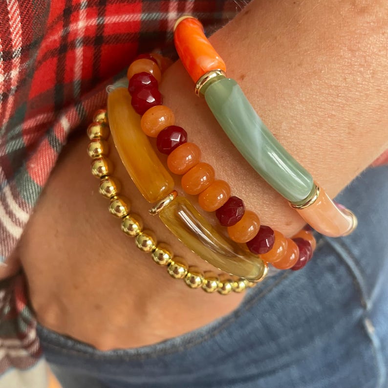 May include: A collection of colorful bracelets. One bracelet features gold beads, while others showcase orange, red, and green beads. The bracelets are stacked on a wrist, with a plaid shirt and denim jeans in the background.