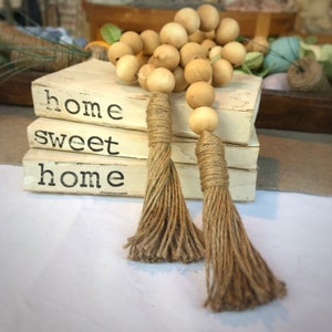 May include: Three white books stacked on top of each other with the words "home sweet home" printed on them. A wooden bead garland with two brown tassels hangs over the books.