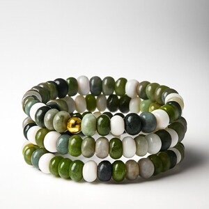 May include: Three stacked bracelets with jade beads in shades of green, white, and black. A gold-colored bead accents the center bracelet. The bracelets are displayed against a white background.