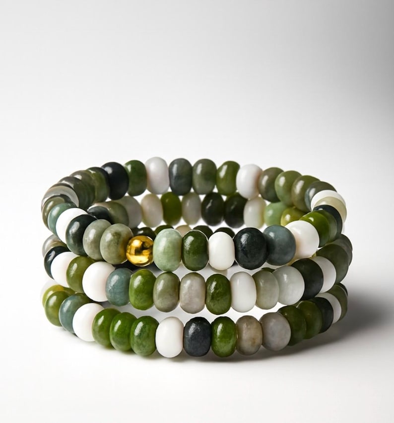 May include: Three stacked bracelets with jade beads in shades of green, white, and black. A gold-colored bead accents the center bracelet. The bracelets are displayed against a white background.