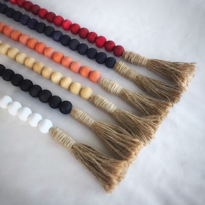 May include: A set of five beaded garlands with tassels. The beads are in shades of red, orange, yellow, beige, and white. The tassels are made of natural jute.
