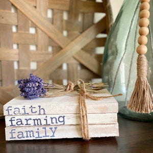 May include: A stack of four white books with the words "faith", "farming", and "family" printed in black on the front. The books are tied together with twine and decorated with a bundle of dried lavender.